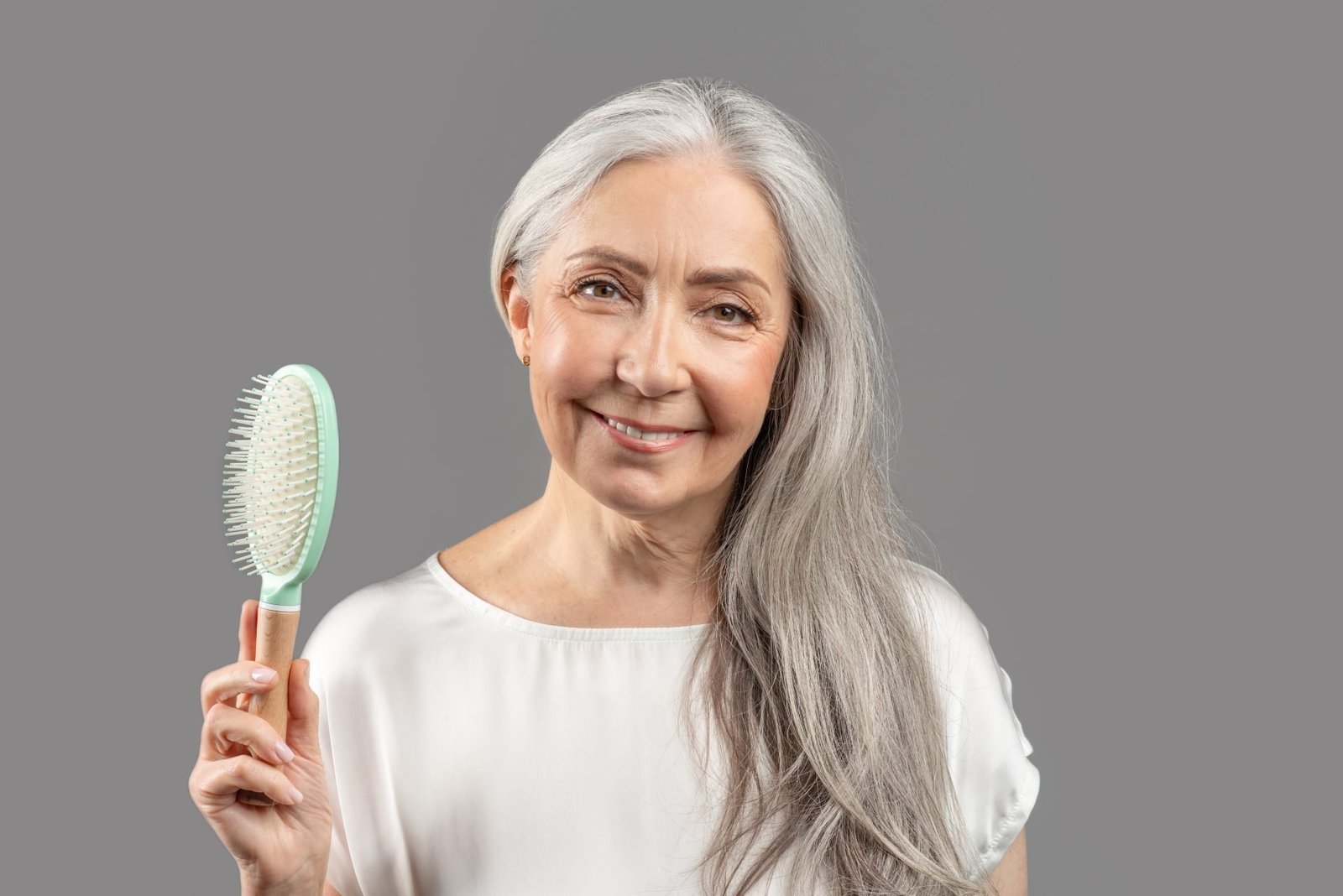 Concetto di COME INFOLTIRE I CAPELLI IN MENOPAUSA. Ritratto di un'affascinante signora anziana con lunghi capelli setosi che tiene in mano una spazzola e sorride su sfondo grigio da studio. Adorabile donna matura che si prende cura di sé.