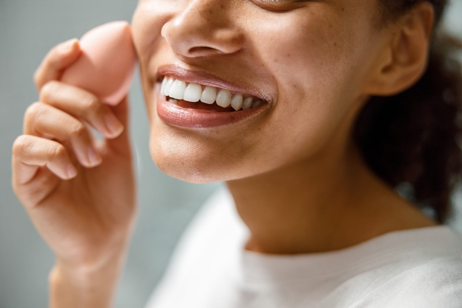 Primo piano di una donna sorridente che si trucca con la skin tint estate 2026 con una spugnetta nel bagno di casa. Foto di alta qualità.