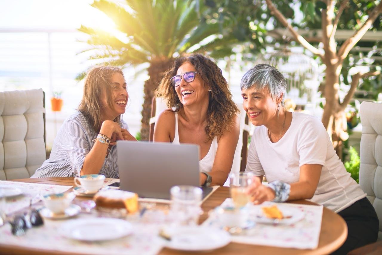 CONCETTO: Come socializzare dopo i 50 anni. Gruppo di donne ride mentre insieme fanno breakfast e guaradno un pc portatile