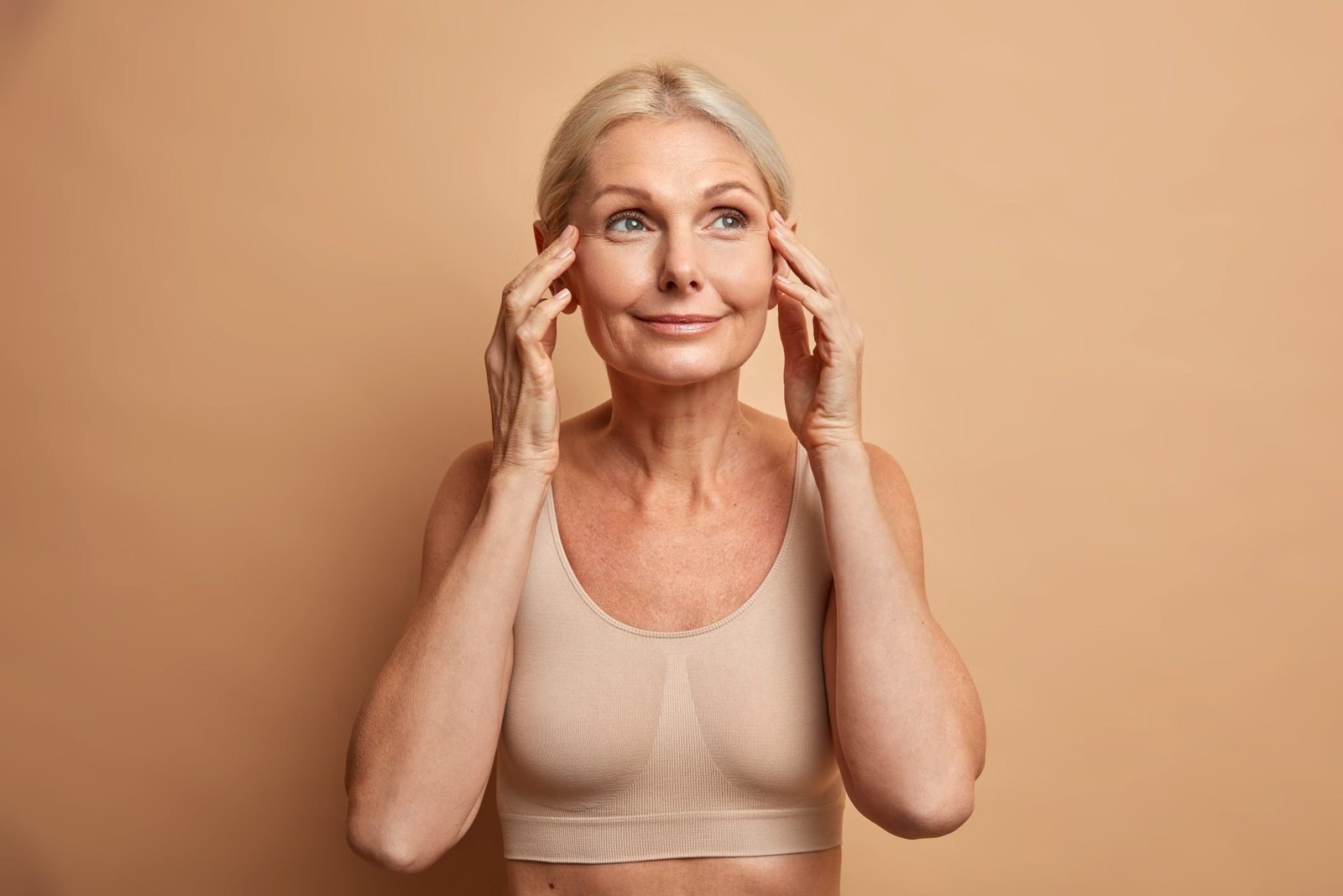 Bellissima donna bionda di mezza età con un sorriso affascinante si tocca il viso e si gode la morbidezza della pelle. Ha un incarnato curato, un trucco naturale e indossa un top corto. concetto di Trucco pelle matura