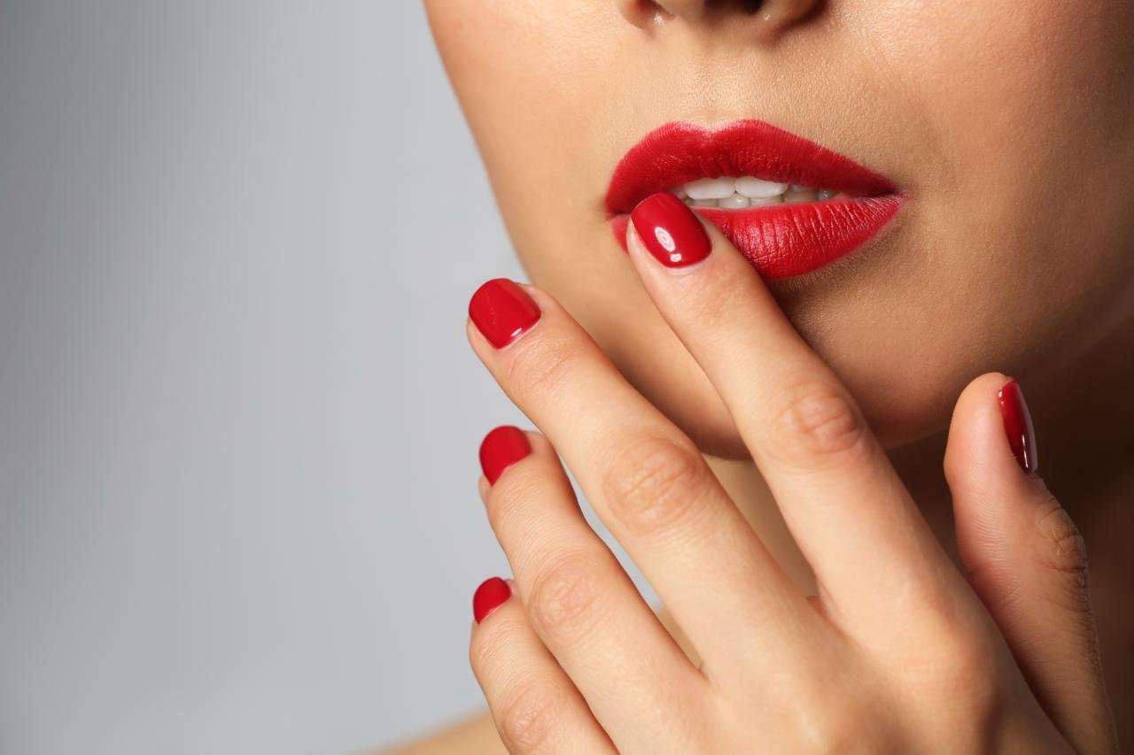 primo piano donna che si tocca con le mani smaltate di rosso, la bocca con rossetto rosso. Tema come far durare smalto a lungo