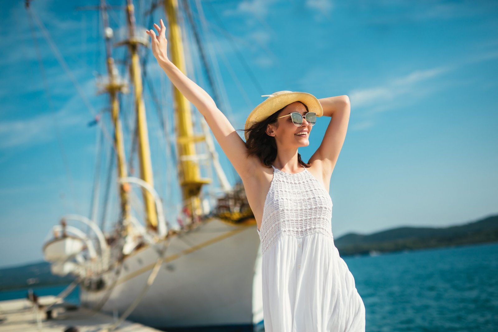 cappelli per il mare : ragazza in abito bianco davanti ad una barca a vela sorride felice in vacanza