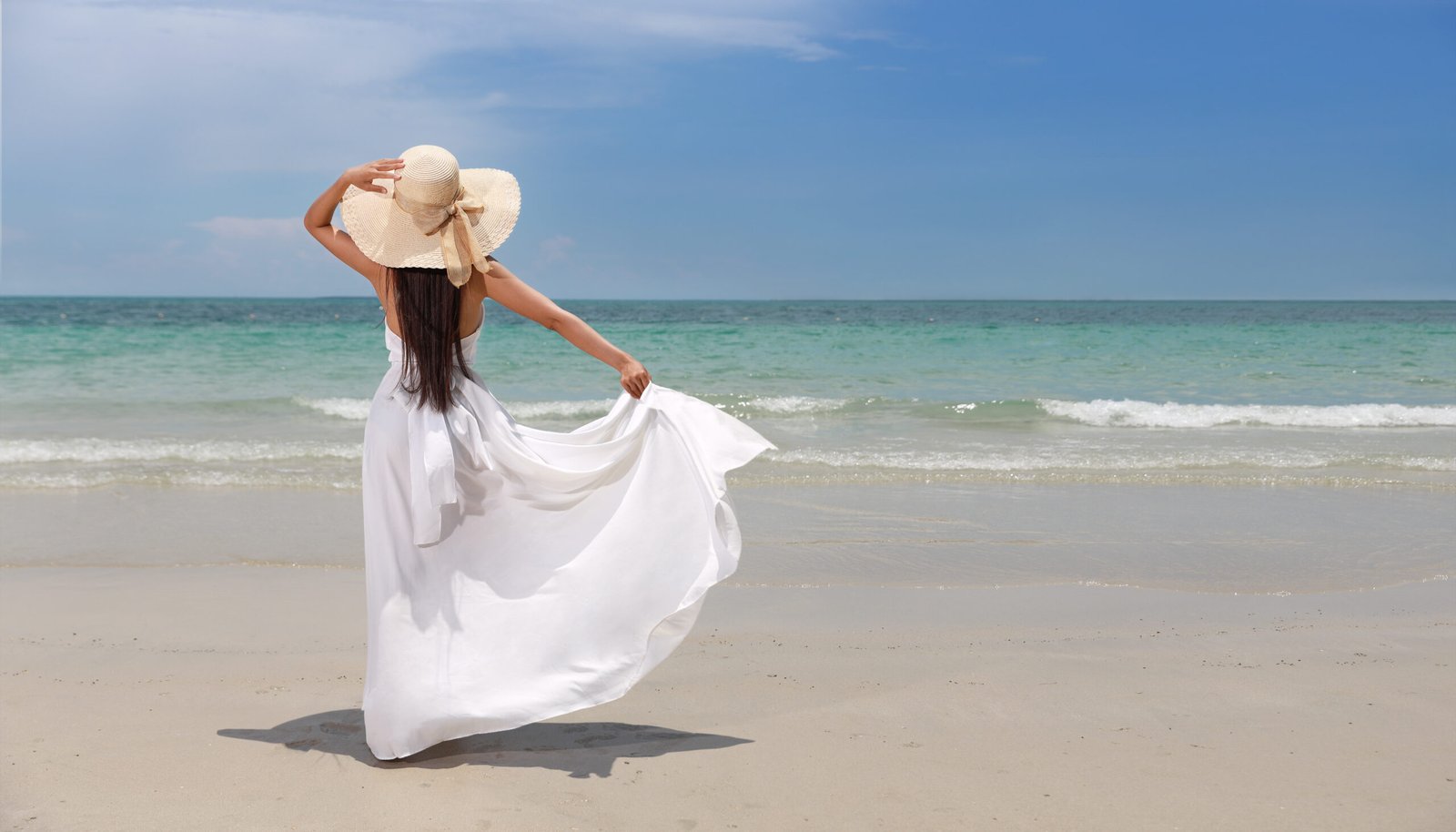 Vista posteriore di una giovane e felice viaggiatrice in abito bianco e cappello in piedi su una bellissima spiaggia sabbiosa.