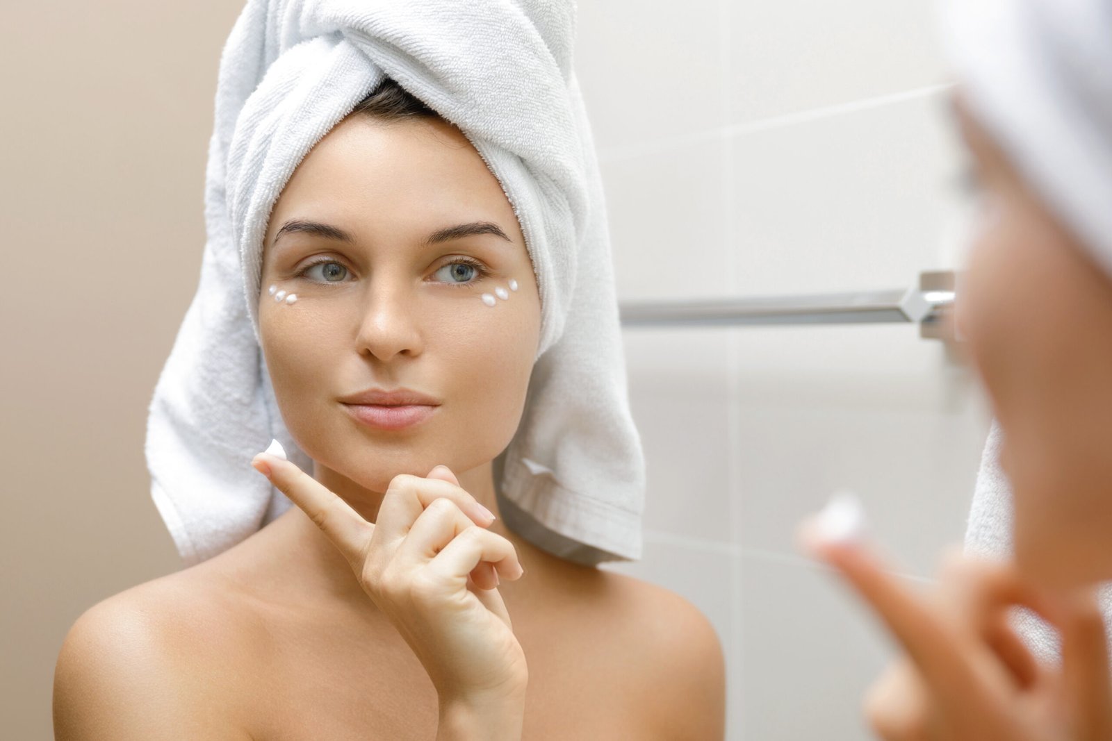 Una donna in bagno si sta applicando nel suo bagno una crema idratante e antietà sotto gli occhi. Ha turbante di spugna in testa. Concetto di routine skincare mare