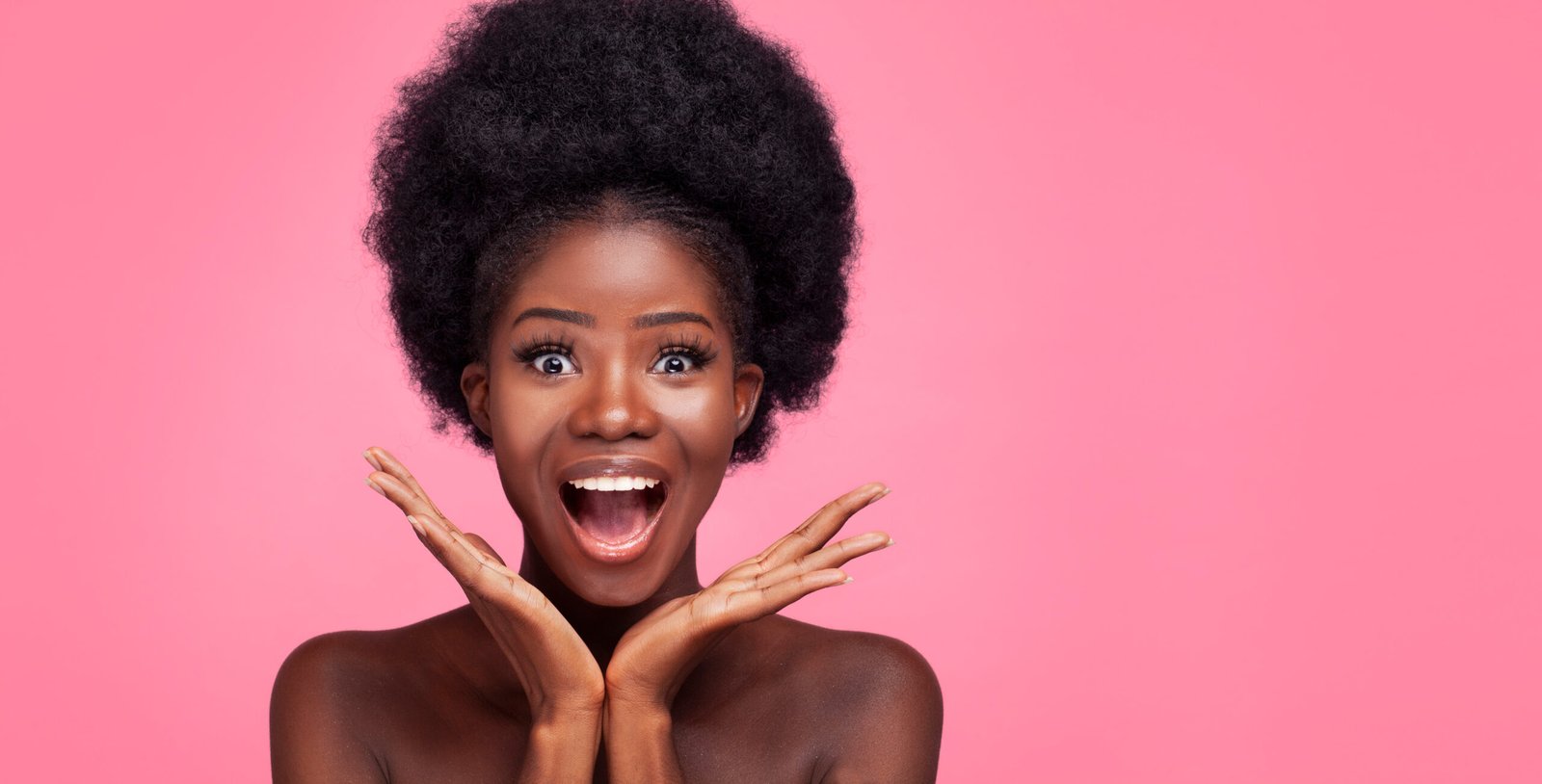 Donna africana sorpresa sorride alla macchina fotografica su sfondo rosa. La giovane ragazza tiene le mani vicino al viso e sorride ampiamente. Concetto di bellezza.