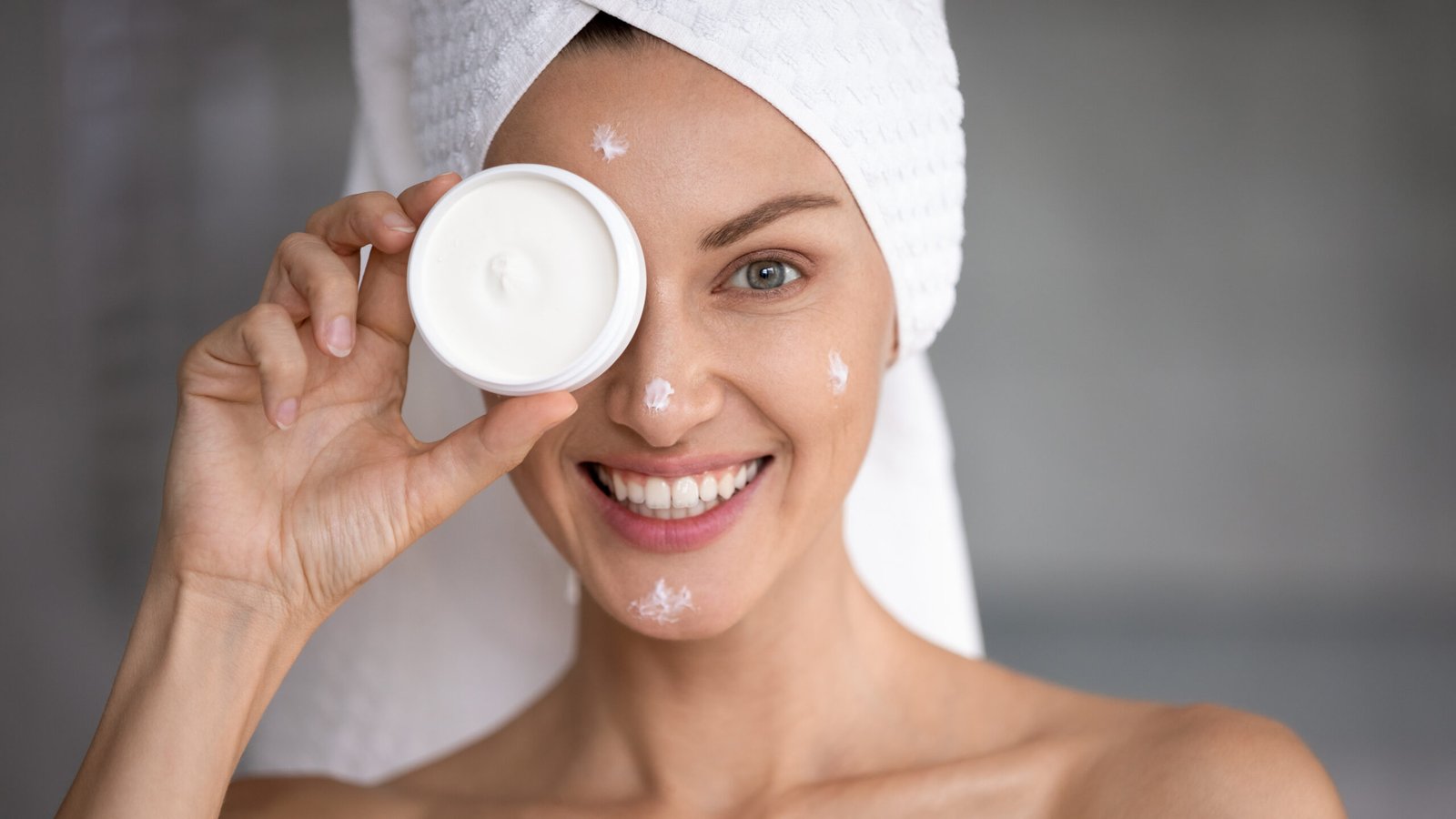 ragazza nel suo bagno con turbante in spugna sulla testa sorride in primo piano e mostra tenendolo davanti al viso un barattolo di crema viso. che ha spalmata in vari punti del viso. Concetto di pelle sana e barriera cutanea protetta