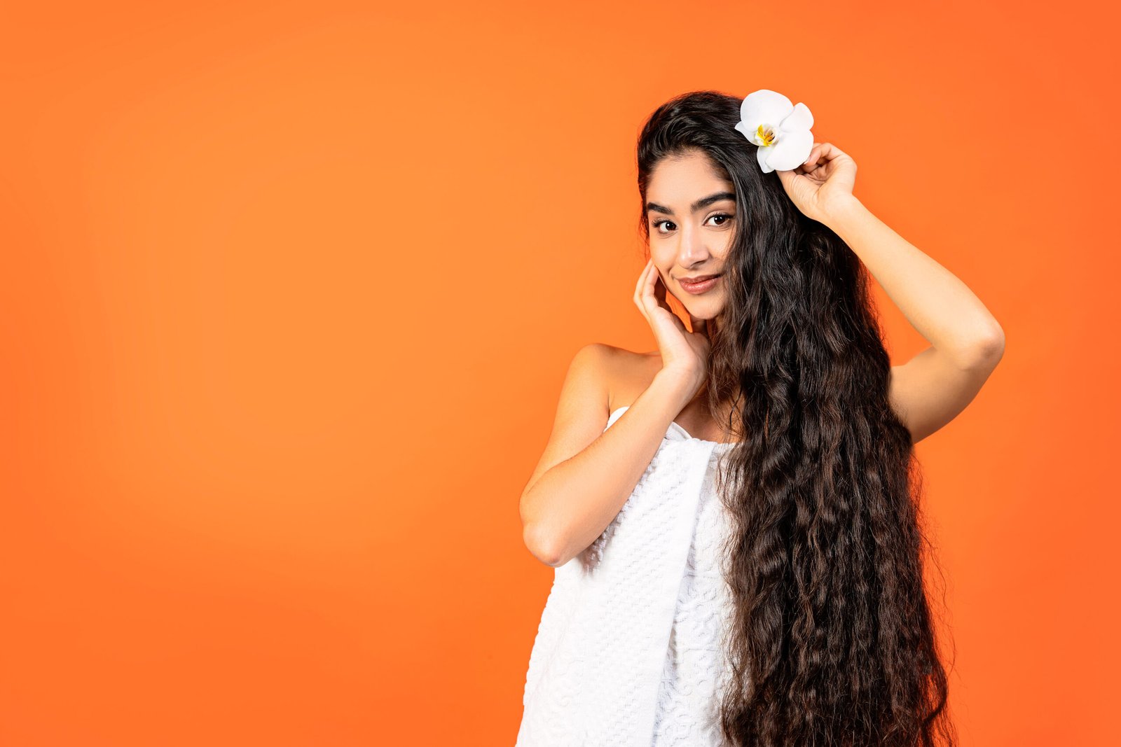 bellissima donna indiana dai lunghi e sani capelli. Indossa un asciugamano bianco, ha un'orchidea tra i capelli e ci guarda. Concetto haircare, cura e integrazione con Biotina per capelli